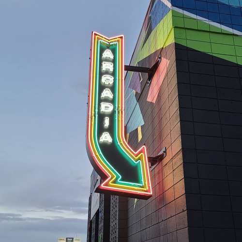 Neon sign for "Arcadia" with an arrow pointing down, mounted on a colorful building exterior.