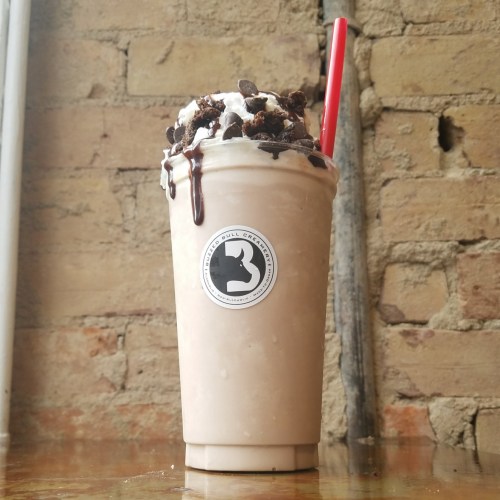 A tall milkshake topped with whipped cream, chocolate drizzle, and crushed cookies, served in a clear cup with a red straw.