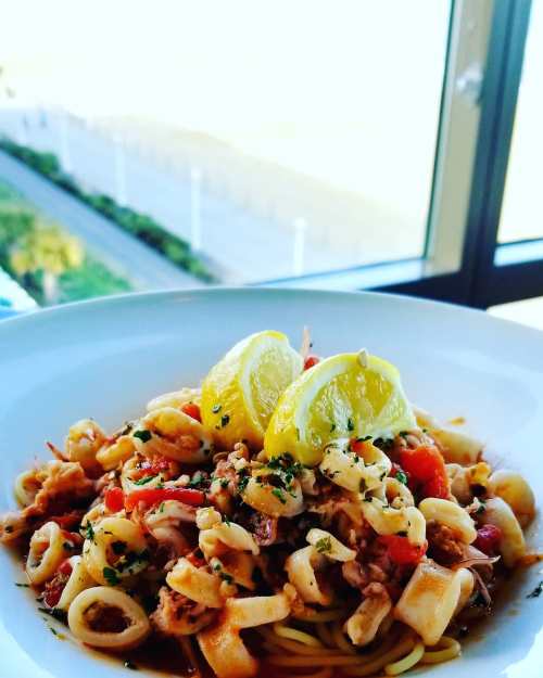A plate of pasta with seafood, topped with two lemon wedges, with a beach view in the background.