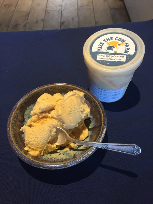 A bowl of caramel ice cream with a spoon, next to a container labeled "Kiss the Cow Farm."