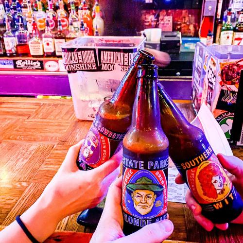 Three hands clink colorful beer bottles together in a lively bar setting, with shelves of liquor in the background.