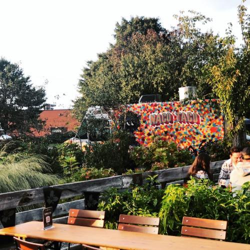 A colorful taco food truck parked in a garden setting with people dining at a wooden table nearby.