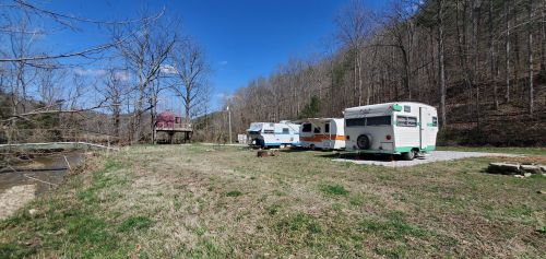 Three vintage RVs parked on a grassy area by a creek, surrounded by trees and a blue sky. A cabin is visible in the background.