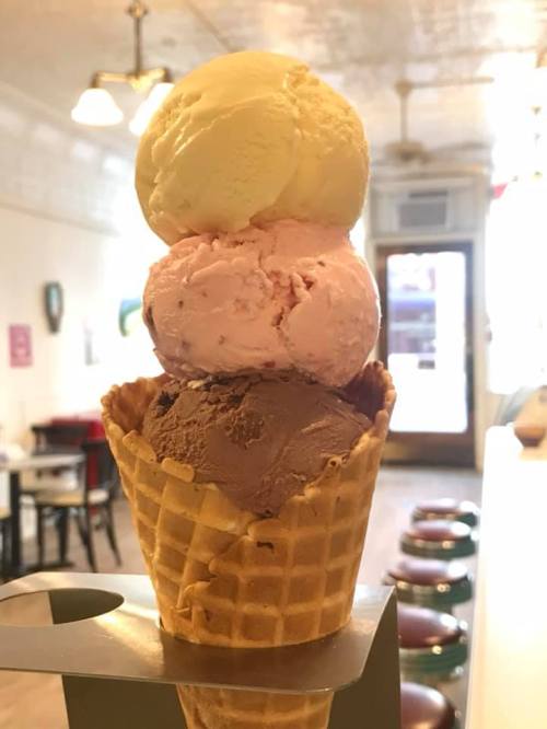 A tall ice cream cone with three scoops: chocolate, pink, and vanilla, set against a cozy ice cream shop background.