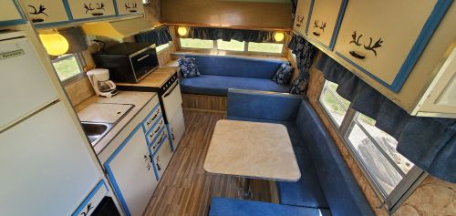 Interior of a vintage camper with blue seating, a small table, kitchenette, and large windows for natural light.