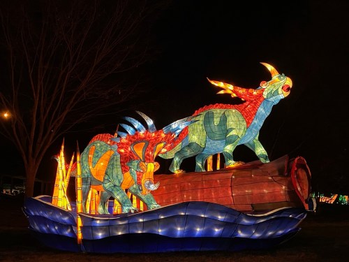 Colorful illuminated lanterns of two dinosaurs on a boat, surrounded by glowing blue waves, set against a dark night sky.