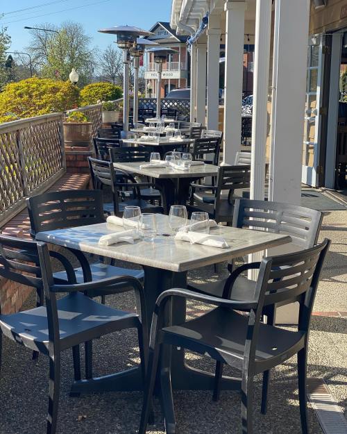 Outdoor dining area with empty tables set for guests, featuring glassware and napkins, surrounded by greenery.