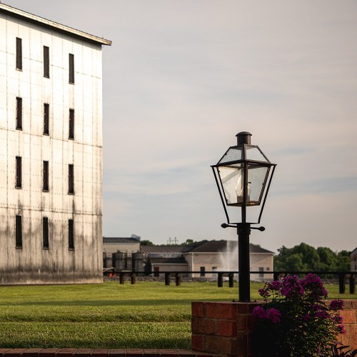 A vintage lamp post stands near a brick wall with purple flowers, beside a large, weathered building in a grassy area.