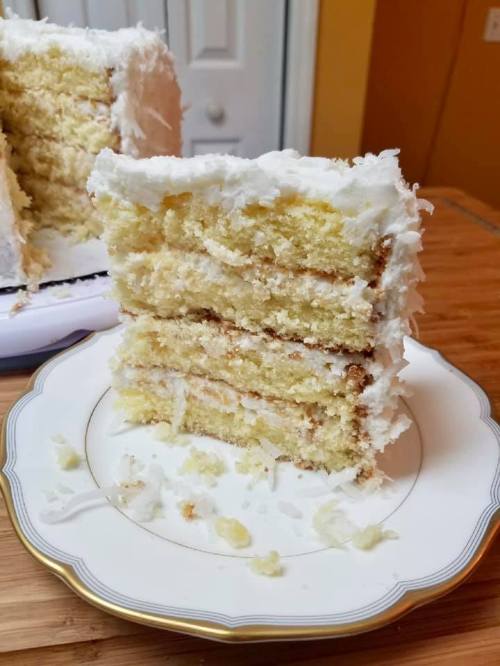 A slice of layered vanilla cake with white frosting on a decorative plate, with the whole cake partially visible in the background.