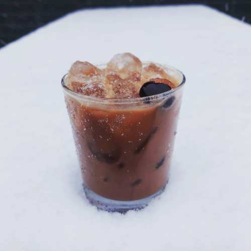 A glass of iced coffee with ice cubes and a cherry, sitting on a snowy surface.