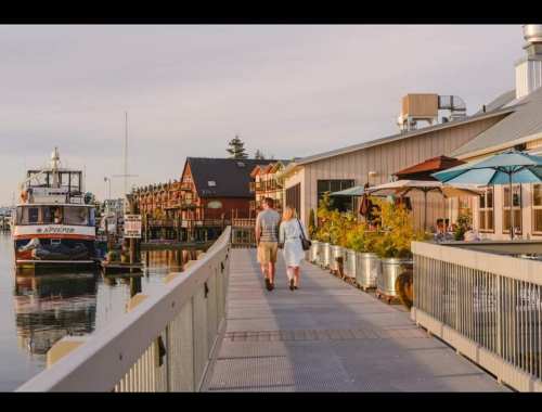 A couple walks along a waterfront pier, with boats and colorful buildings in the background.
