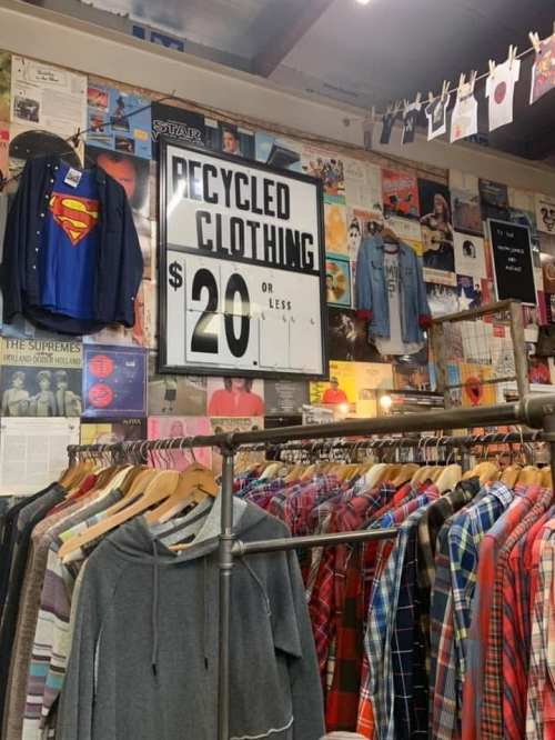 A thrift store display featuring a sign for recycled clothing priced at $20 or less, with various shirts hanging nearby.