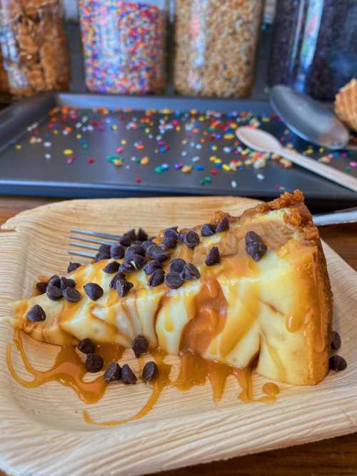 A slice of cheesecake drizzled with caramel and topped with chocolate chips, served on a wooden plate.