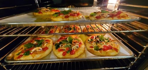 Mini pizzas topped with sauce, vegetables, and herbs baking in an oven on two trays.