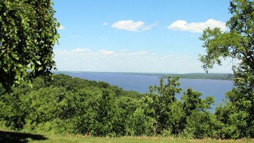 A serene view of a river surrounded by lush green trees under a clear blue sky with scattered clouds.