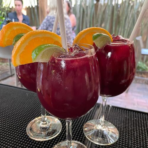 Three colorful cocktails with orange and lime garnishes, served in large glasses on a bar counter.