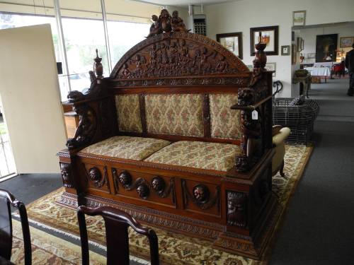 Ornate wooden bench with intricate carvings and patterned upholstery, displayed in an art gallery setting.