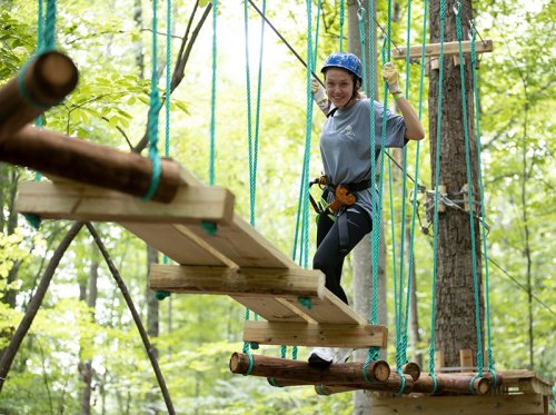 A young person navigates a wooden rope bridge in a forested adventure park, wearing a helmet and harness.
