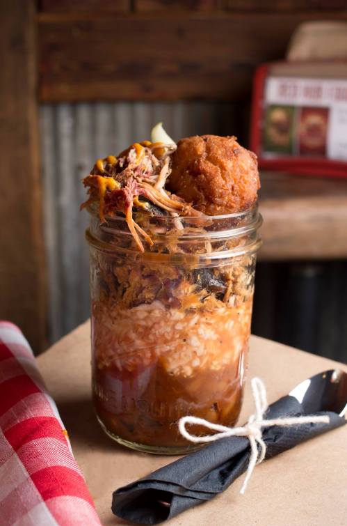 A mason jar filled with pulled pork, rice, and a cornbread muffin on top, set on a rustic table with a checkered napkin.