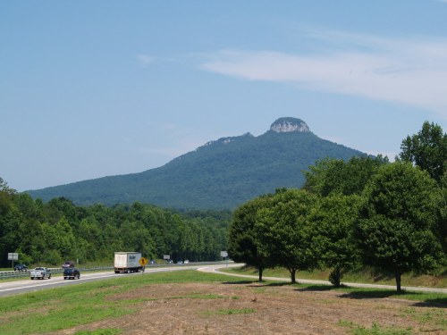 A scenic view of a mountain with a flat top, surrounded by trees and a highway in the foreground.