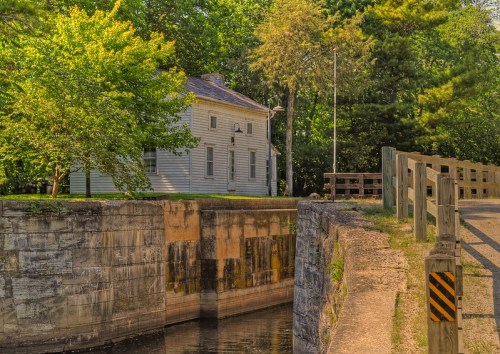 A historic building beside a canal, surrounded by trees, with a stone wall and wooden railing in the foreground.