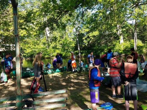 A group of people in life jackets gather by a pond, surrounded by trees, preparing for an outdoor activity.