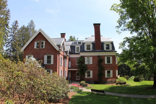 A large red brick house with multiple chimneys, surrounded by greenery and colorful flowers on a sunny day.