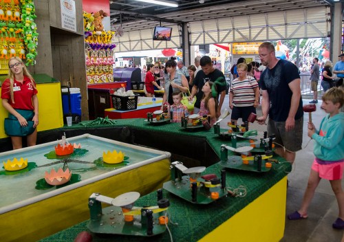 A busy carnival game area with families playing, colorful prizes, and a water lily-themed game setup.
