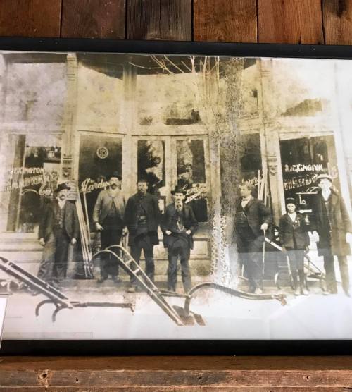 Black and white photo of seven men standing outside a storefront, with vintage signage visible in the background.