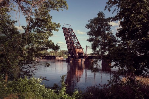 A rusted drawbridge partially raised over a calm river, surrounded by lush greenery and trees under a clear sky.