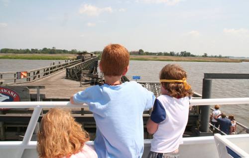 Three children on a boat, looking out at a wooden pier and calm water on a sunny day.
