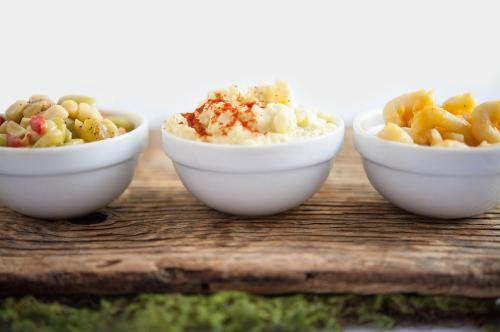 Three small white bowls on a wooden board, filled with different dishes: a salad, creamy dip, and pasta.