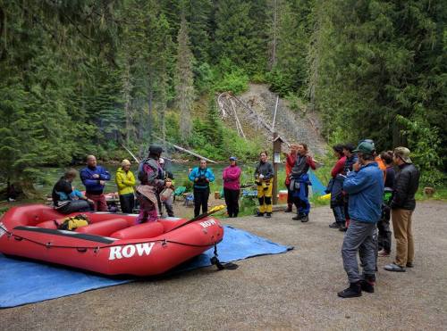 A group of people gathers around a red raft in a forested area, listening to a guide near a river.