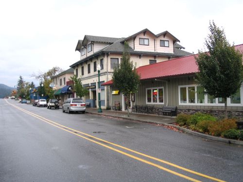 A quiet street scene featuring shops and buildings with a cloudy sky overhead. Cars are parked along the road.