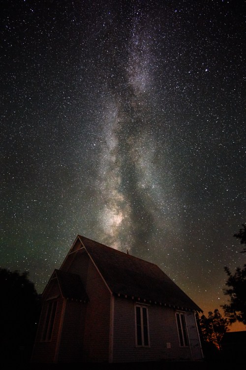 A starry night sky filled with the Milky Way above a small, silhouetted house.