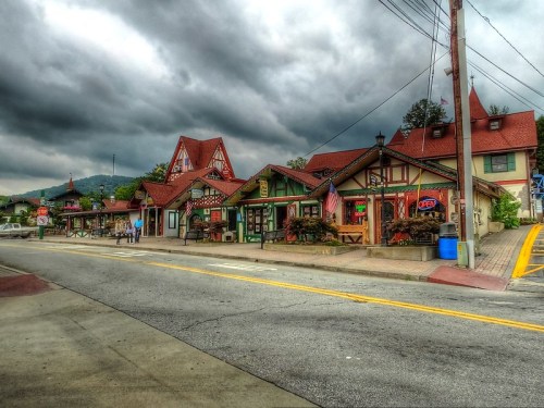Charming alpine-style buildings line a street under a cloudy sky, with shops displaying colorful signs and decorations.