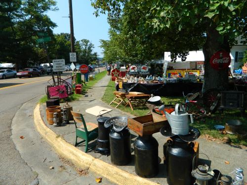 A roadside market featuring vintage items, including metal containers, furniture, and signs, under a large tree.