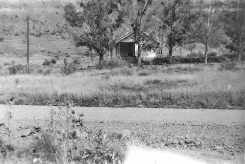 A black and white photo of a small house surrounded by trees, set back from a gravel road in a rural landscape.