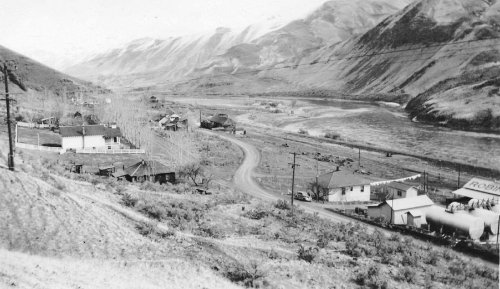 Black and white landscape of a valley with a winding road, river, and small buildings surrounded by mountains.