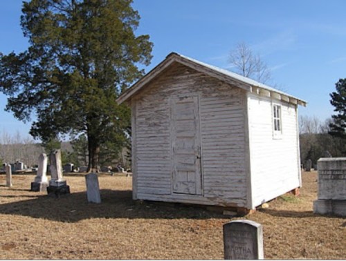 A small, weathered white shed stands in a cemetery, surrounded by gravestones and trees under a clear blue sky.