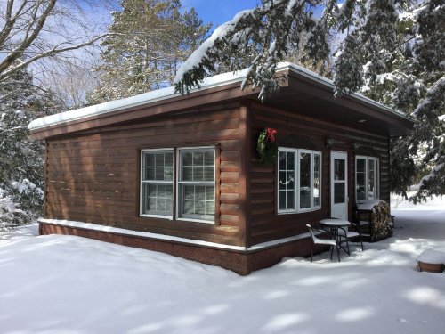 A cozy log cabin covered in snow, with a wreath on the door and surrounded by trees under a clear blue sky.