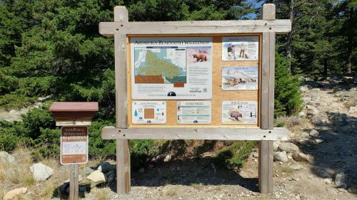 Information sign at a trailhead, featuring maps and wildlife safety tips, surrounded by trees and rocky terrain.