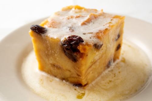 A square piece of bread pudding topped with a creamy sauce and dotted with raisins, served on a white plate.