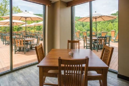 A wooden dining table with chairs, overlooking a patio with umbrellas and greenery outside.