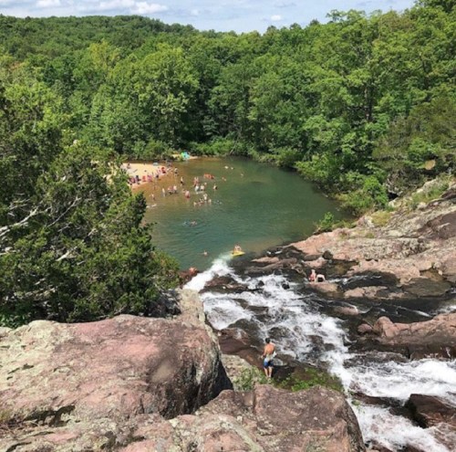 A scenic view of a rocky waterfall leading to a swimming area surrounded by lush green trees and people enjoying the water.