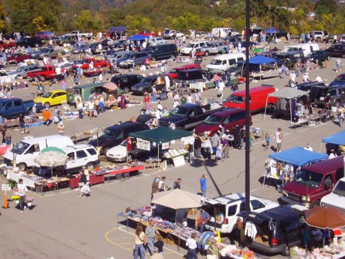 A bustling outdoor market with numerous cars and vendors selling various goods under colorful tents.