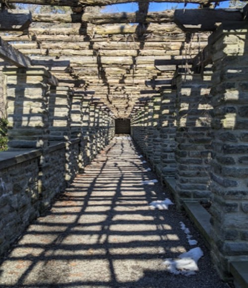 A stone pathway lined with columns, covered by a wooden trellis, casting intricate shadows on the ground.