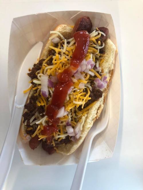 A loaded hot dog topped with chili, shredded cheese, diced onions, and ketchup, served in a paper tray with utensils.