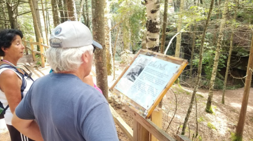 Two people observe a sign in a forested area, with trees and a rocky background visible.