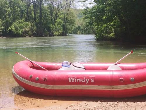 A red inflatable raft named "Windy's" rests on a sandy riverbank surrounded by lush green trees.